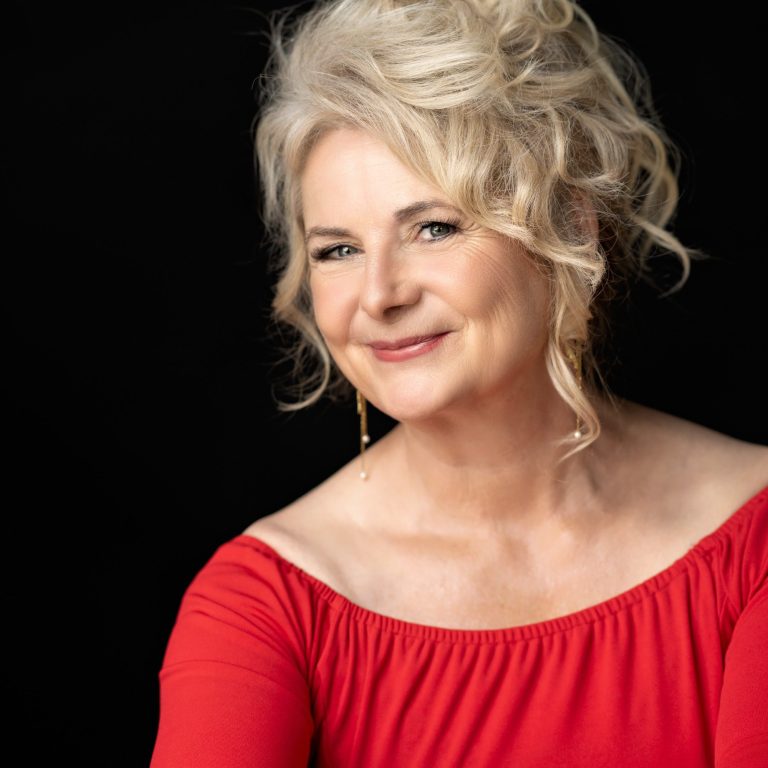 Frau mit blonden Locken, rotem Oberteil und freundlichem Lächeln, vor schwarzem Hintergrund.