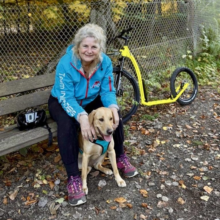 Eine Frau sitzt auf einer Bank mit einem Hund, neben ihr steht ein gelbes Tretboard.